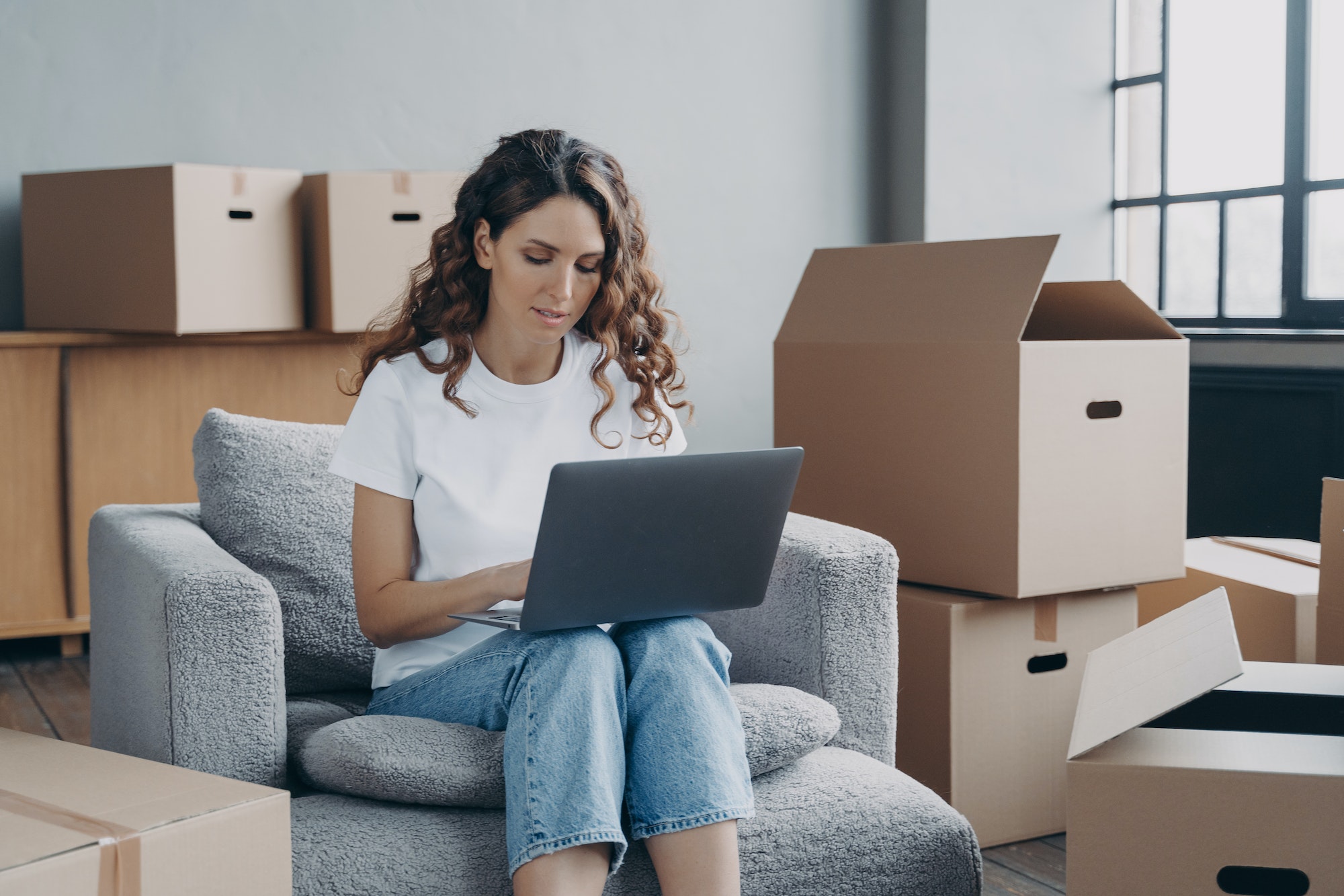 Girl shopping online for new house at laptop, sitting among card boxes. Relocation, ecommerce